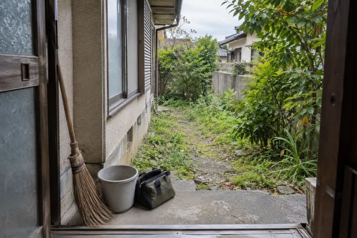 玄関のほうき、バケツ、道具袋と、外壁沿いの通路や草木を描いた生成画像