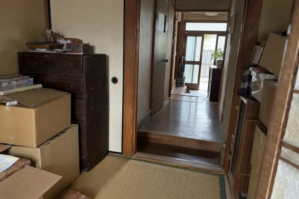 古い日本の住宅の部屋から玄関を見る説明用生成写真。箱とたんすが残り、通路に段差と狭い出入口が見える
