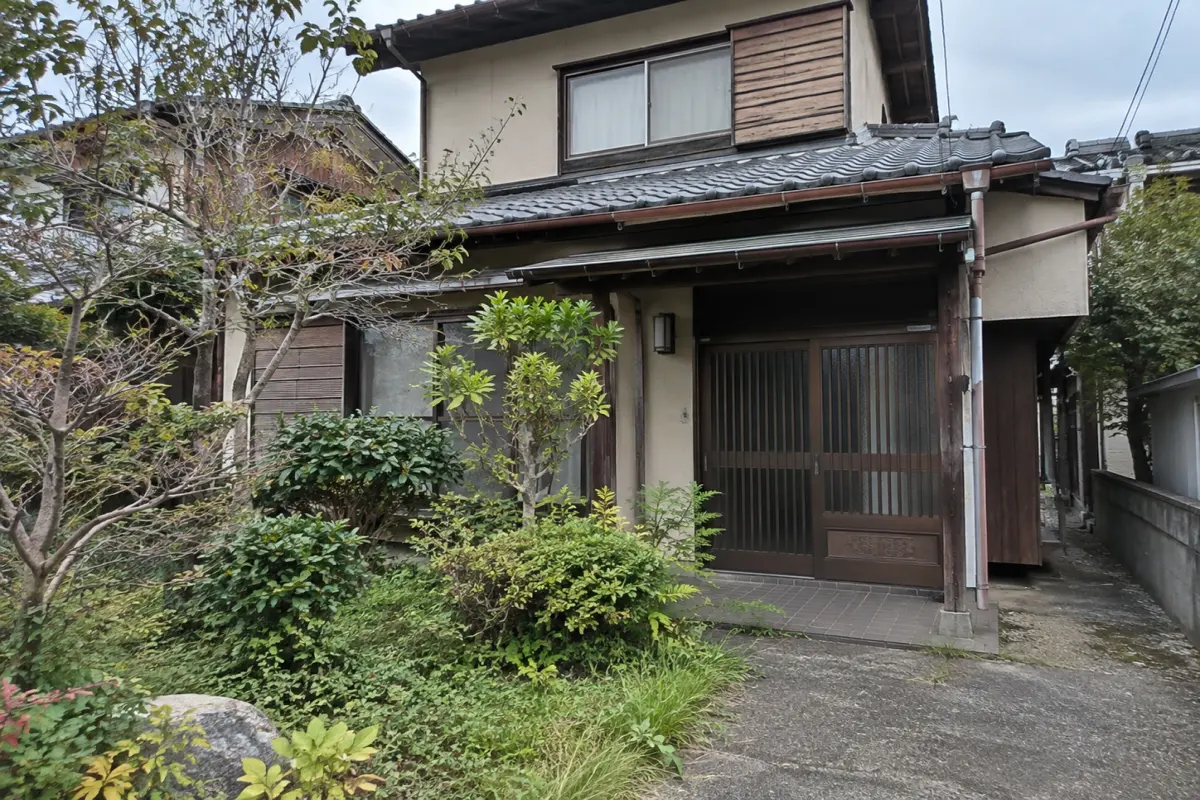 植木と外壁、玄関前の通路が見える古い日本の戸建て住宅の説明用生成写真