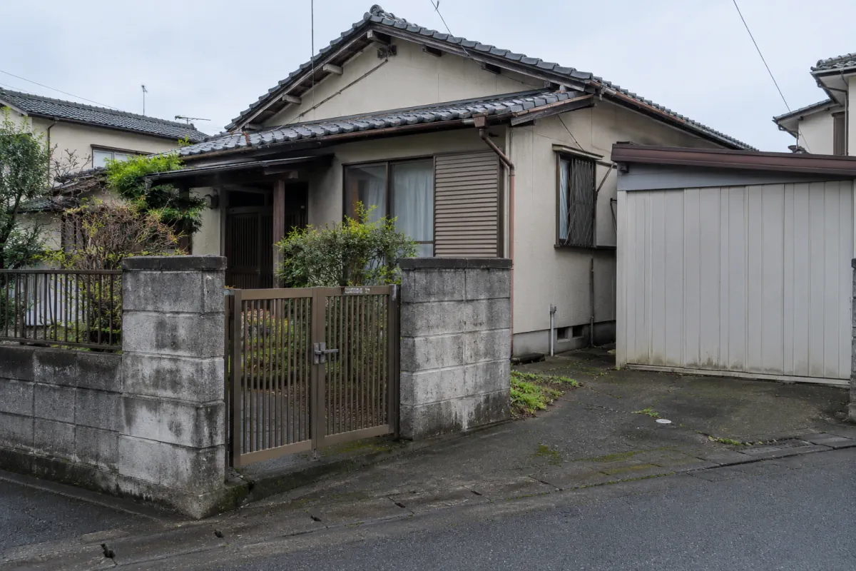 古い日本の住宅を道路から写した説明用生成写真。母屋、脇の小さな車庫、門と塀が見える