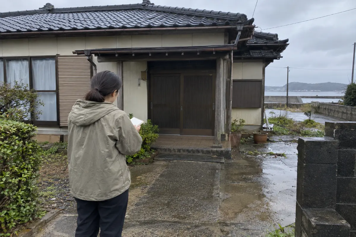 雨上がりの古い家の入口を、ノートを持つ人が外から確認する生成イメージ