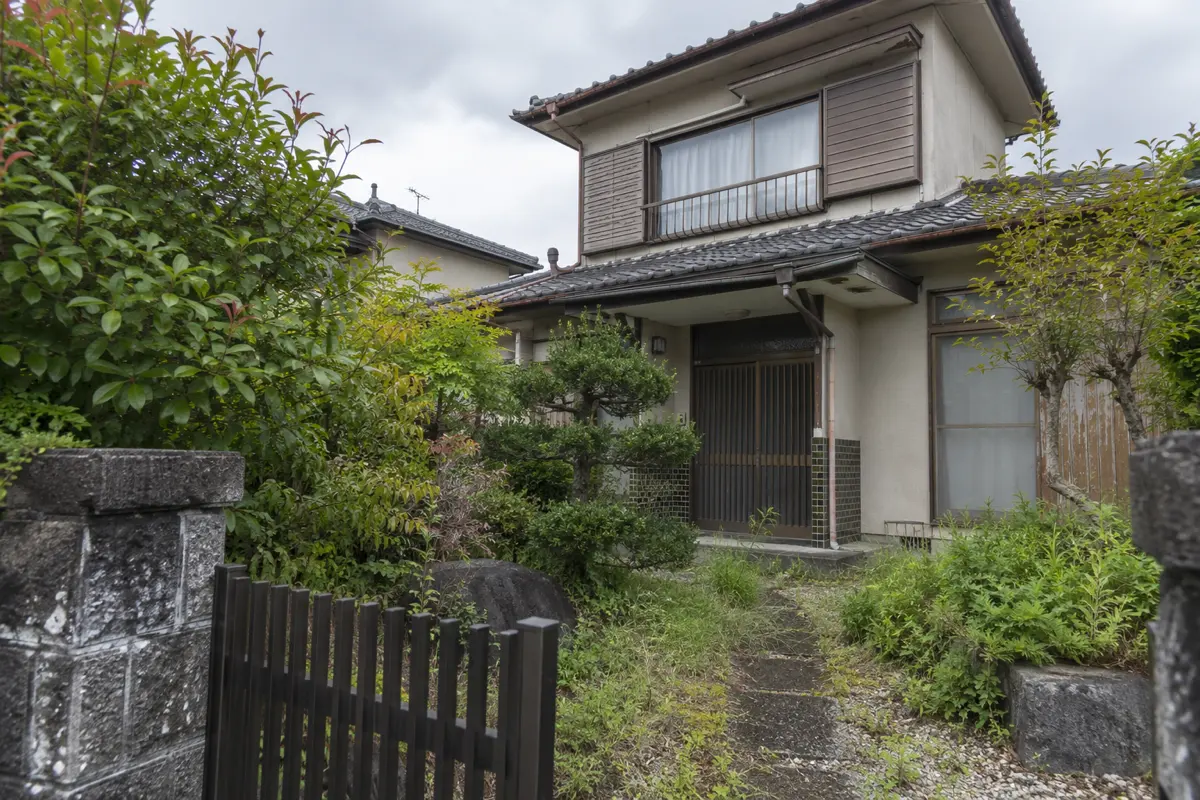 庭木が伸びた古い日本の戸建て住宅を門の外から見た説明用生成写真